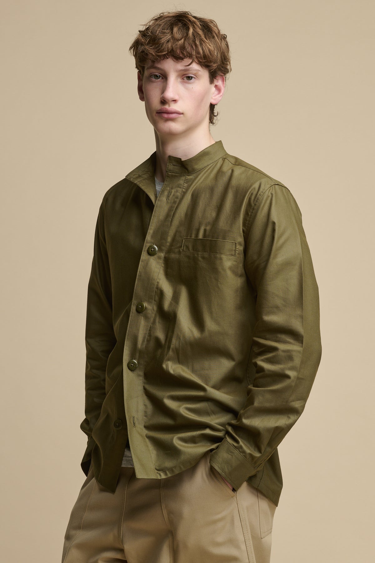Male wearing men's George Lightweight Collarless Overshirt in Olive worn with half unbuttoned over grey t shirt paired with Cameraman pants in putty