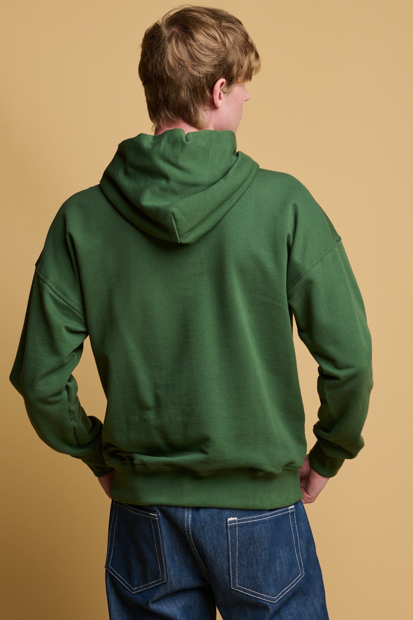 Thigh up image of the back of male wearing hooded sweatshirt in bottle green paired with community clothing blue arran button fly jeans