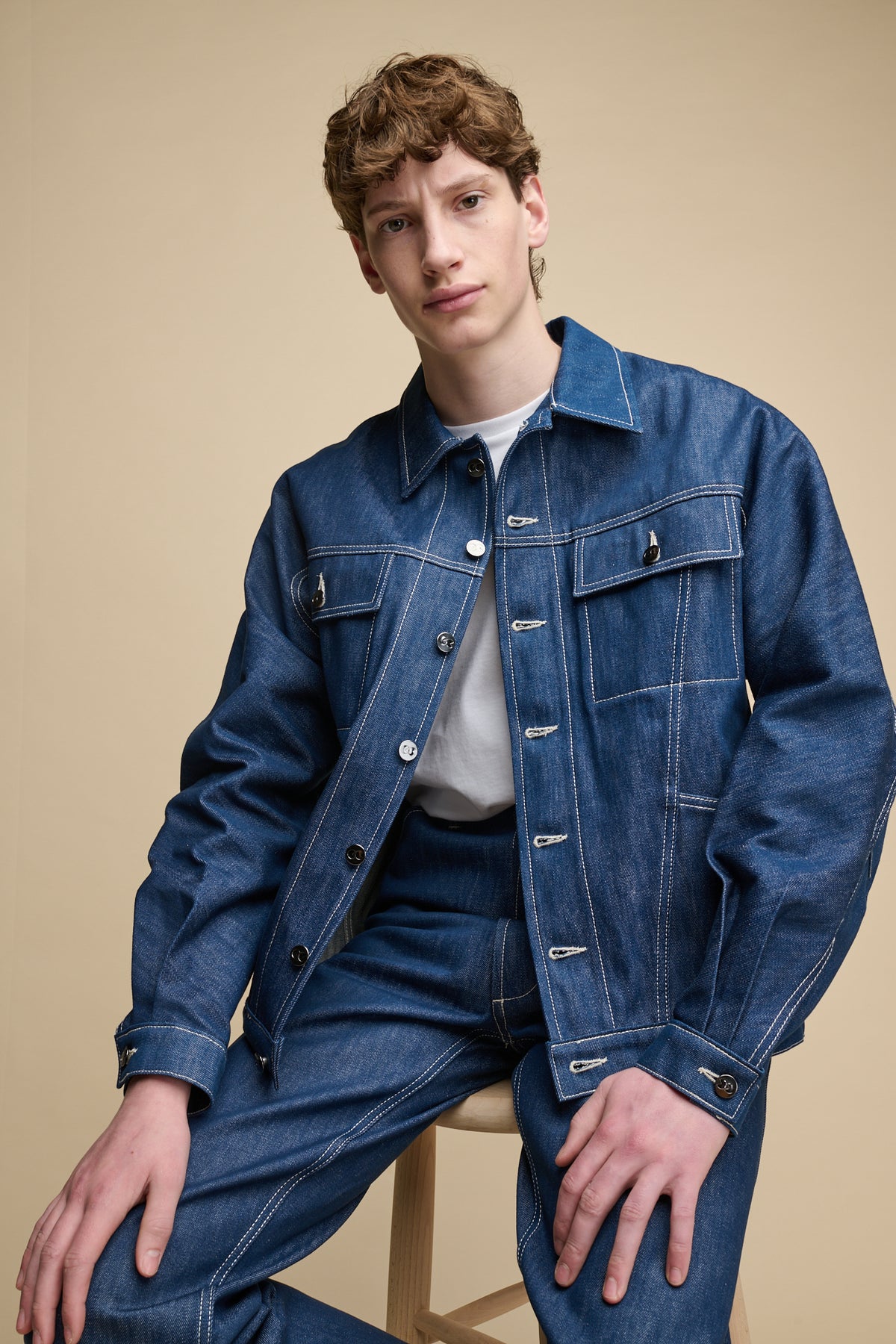 Knee up image of male sat on stool wearing Joe Denim Jacket in Blue with matching denim jeans 