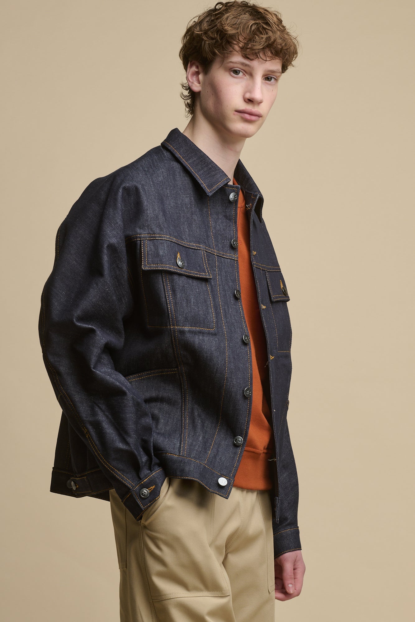 Thigh up image of male standing on an angle looking towards the camera wearing Joe Denim Jacket in indigo worn with raglan sweatshirt in cinnamon and cameraman pants in putty