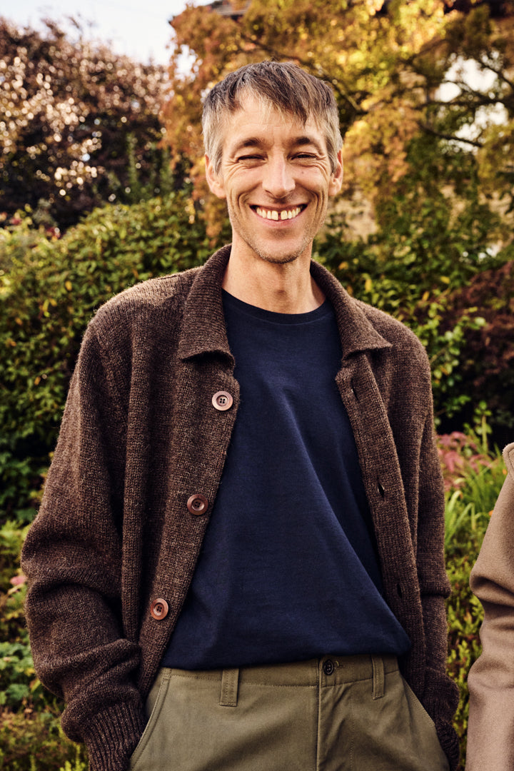 Man wearing a brown cardigan over a navy shirt with green pants, standing outdoors.