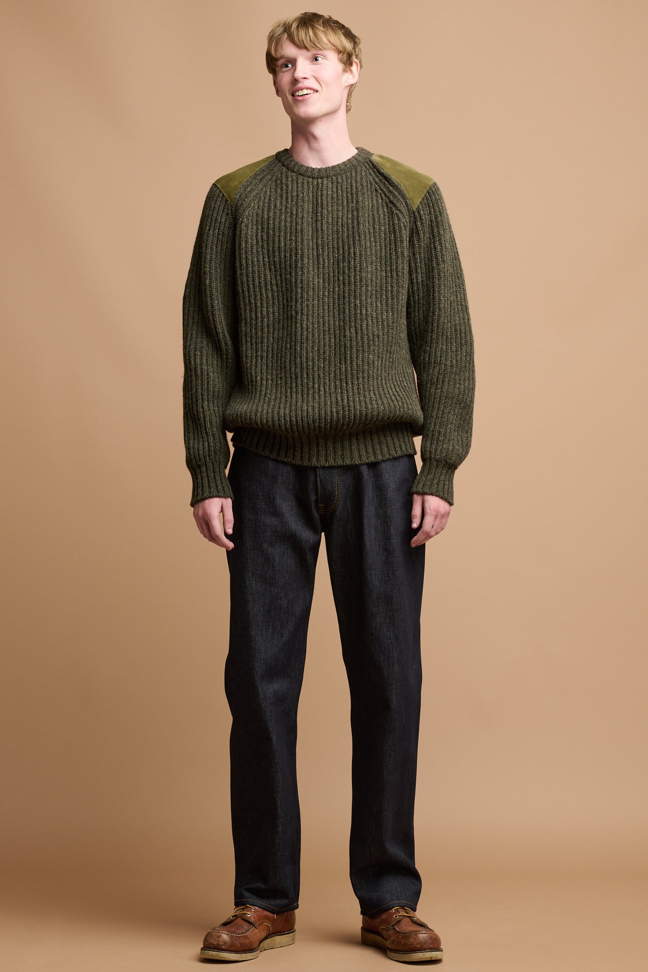 The front of male wearing lambswool fisherman ribbed jumper in dark olive paired with loose fit button fly arran jean in indigo