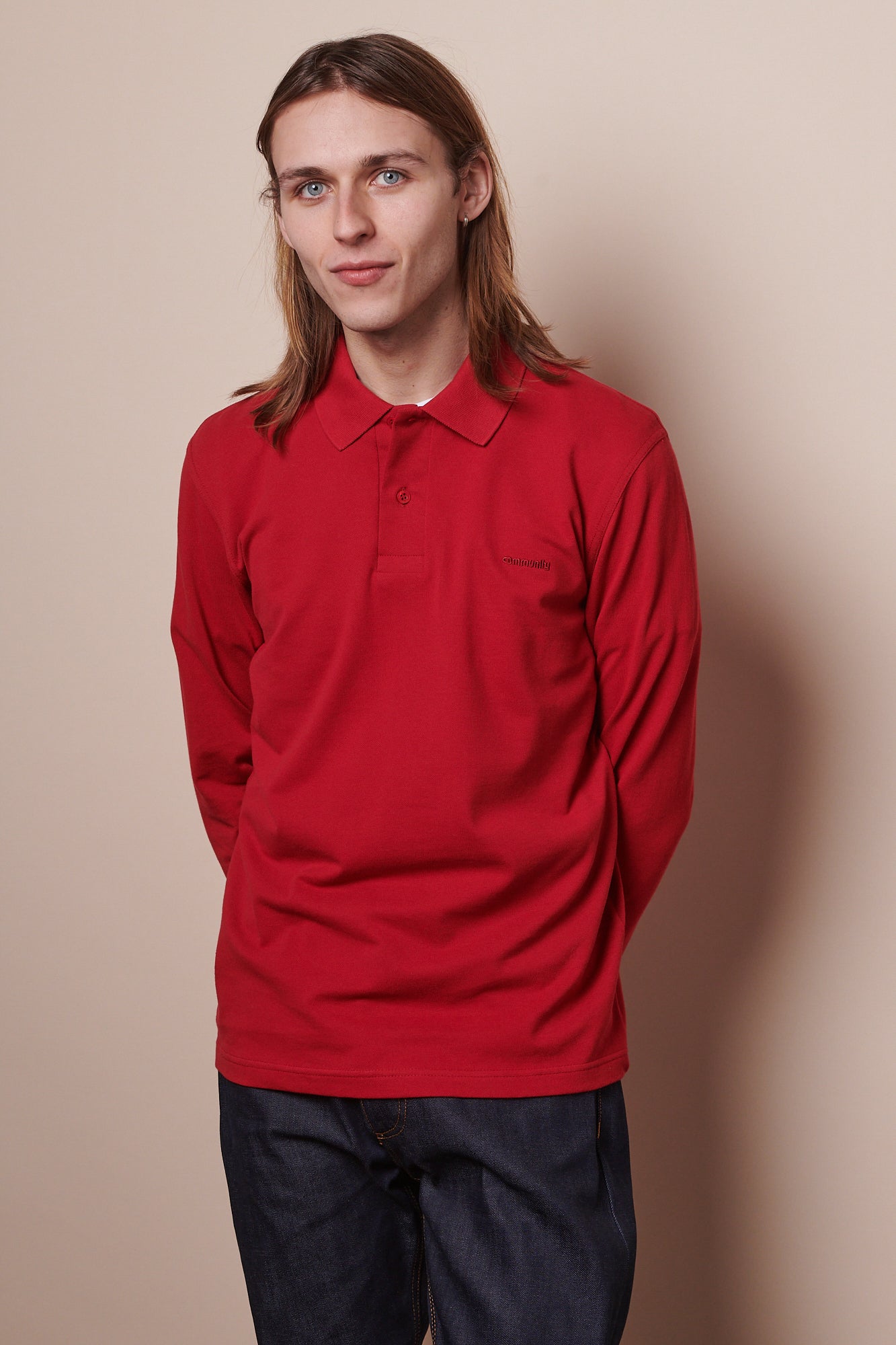 Thigh up image of white male with shoulder length brown hair wearing long sleeve t shirt in crimson with arms held behind back 