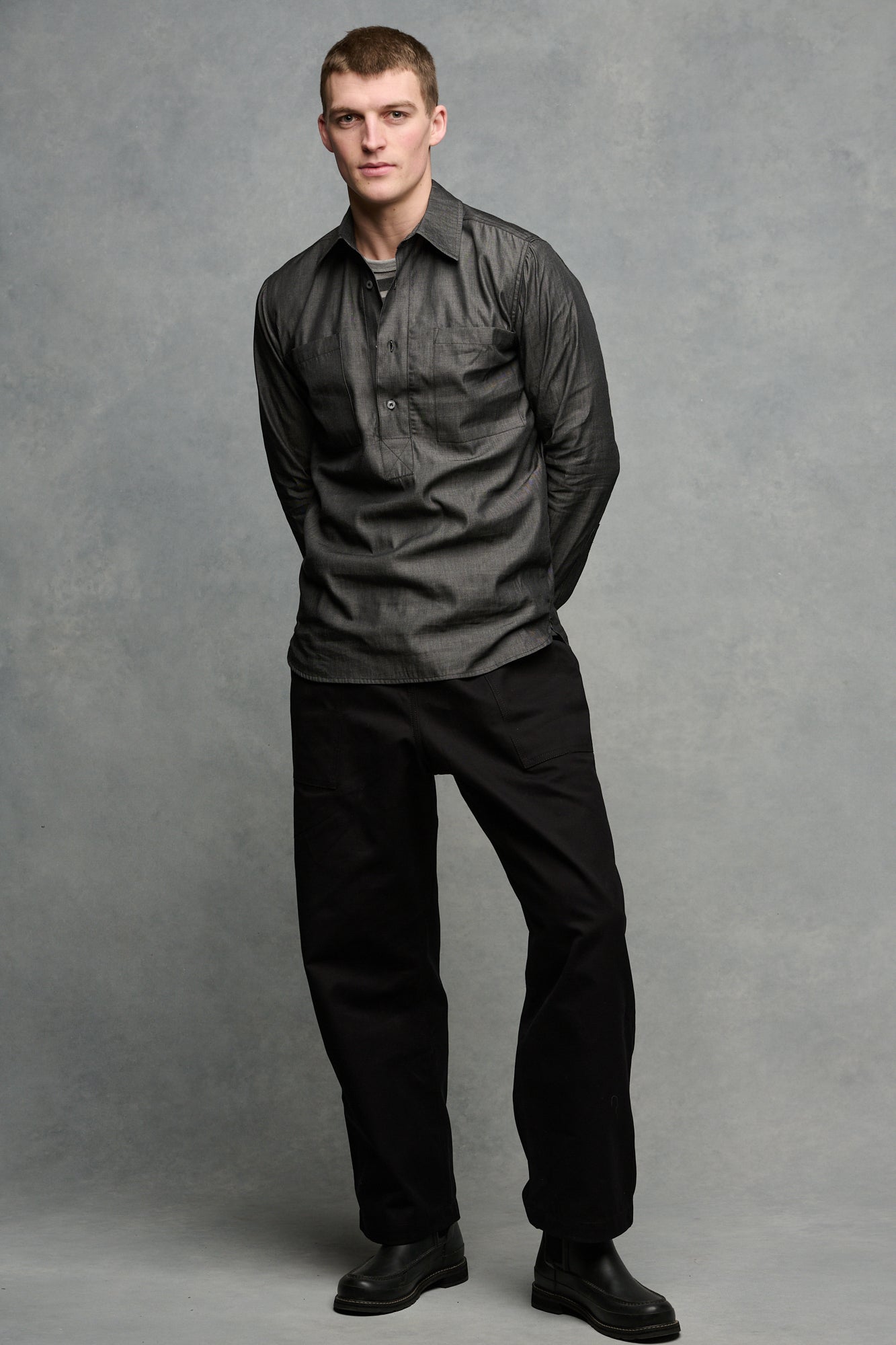 Full body image of male wearing Oli half placket lightweight denim shirt in black paired with his hands behind the back paired with cameraman pants in black and black boots