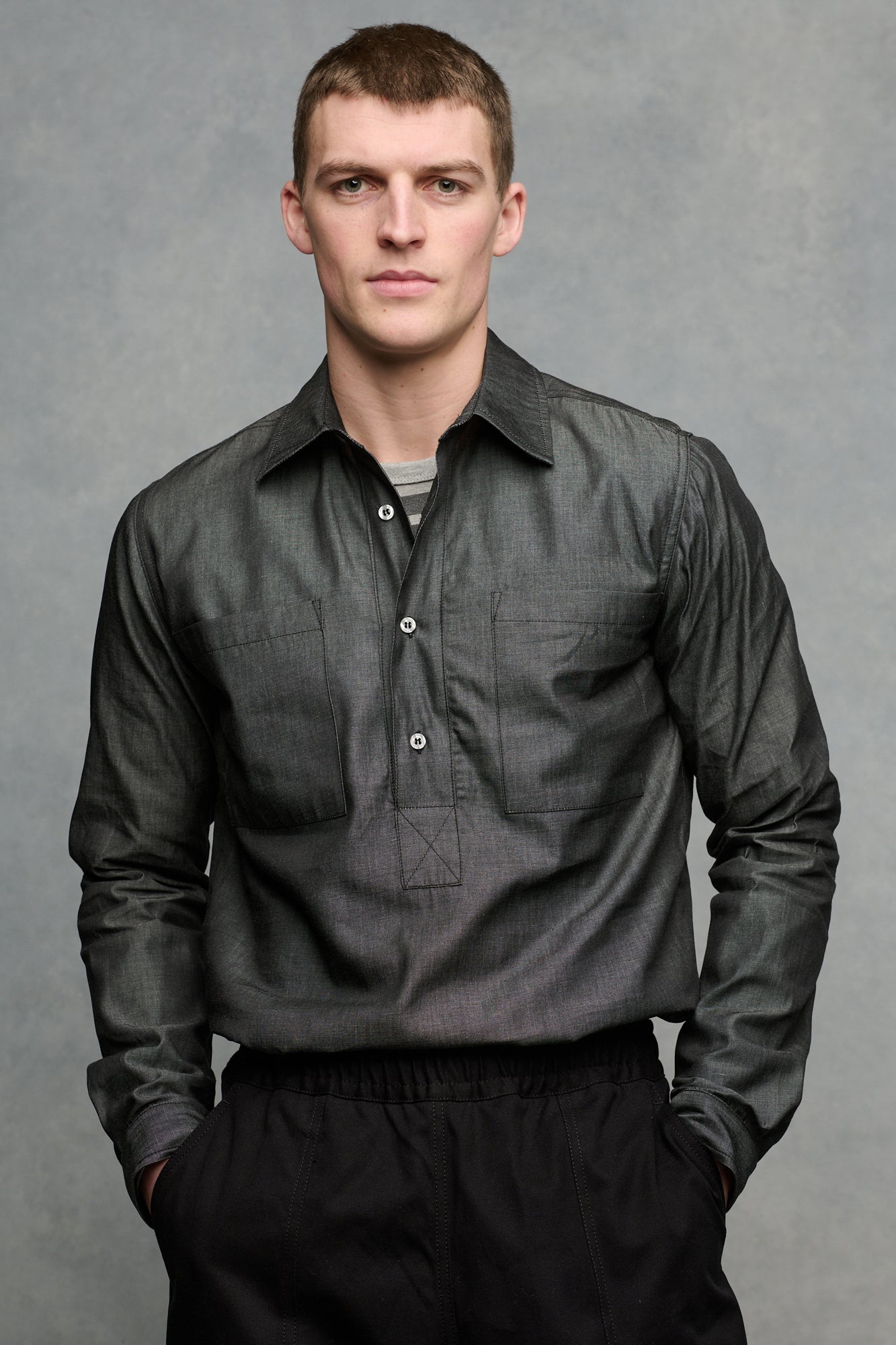 Thigh up front of male wearing Oli half placket lightweight denim shirt in black tucked into cameraman pants in black