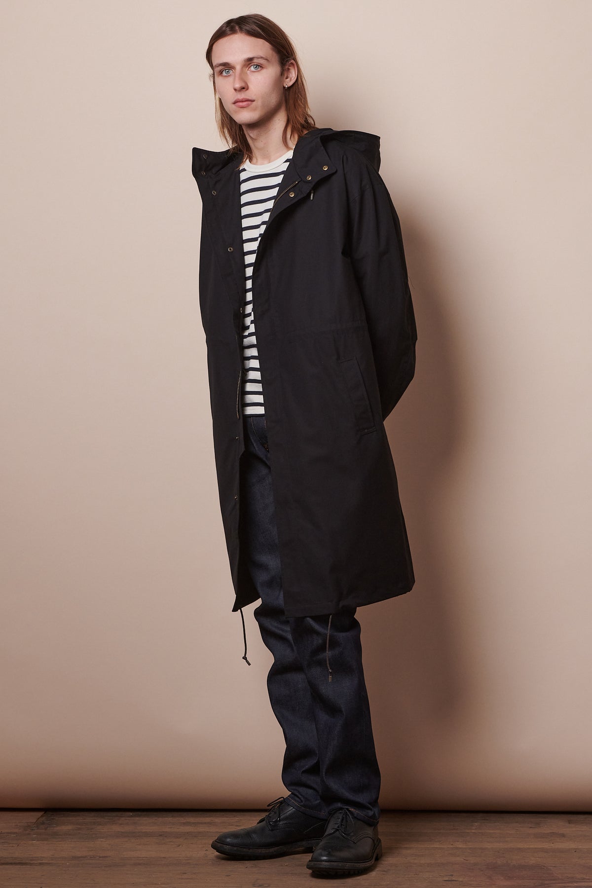 Full body image of male wearing black parka unzipped holding hands behind back.