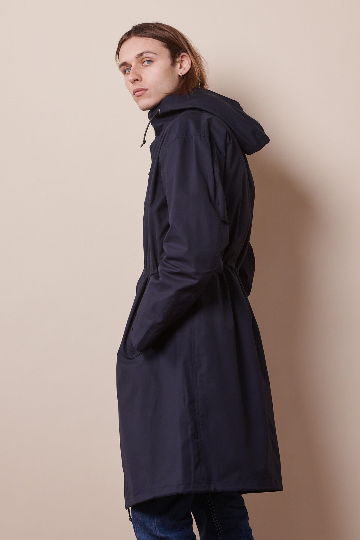Back of male looking back towards camera wearing navy parka with hands in pockets. Adjustable waist cord tightened to pull waist in 