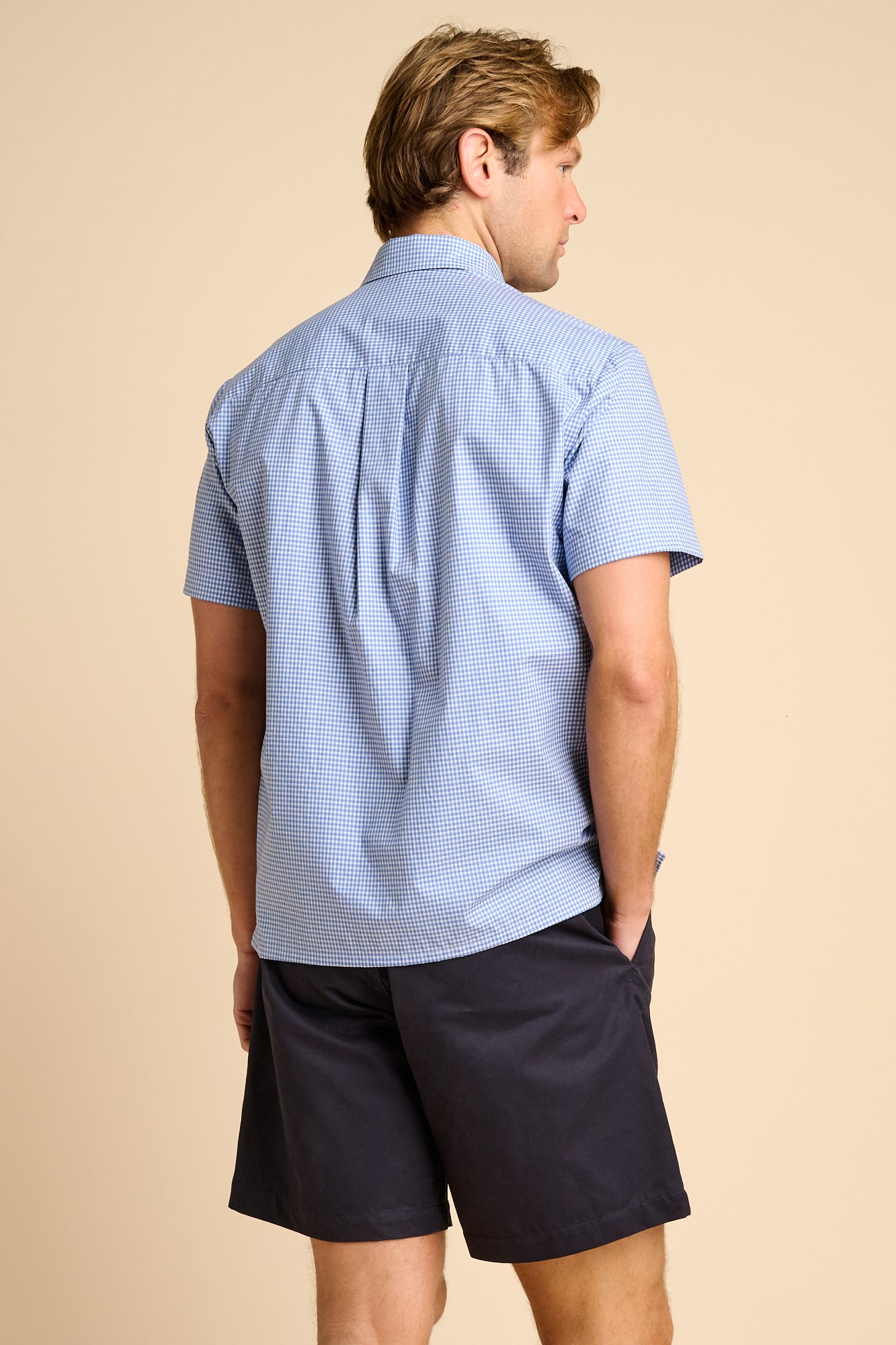 Man wearing a light blue checkered shirt and dark shorts on a beige background.