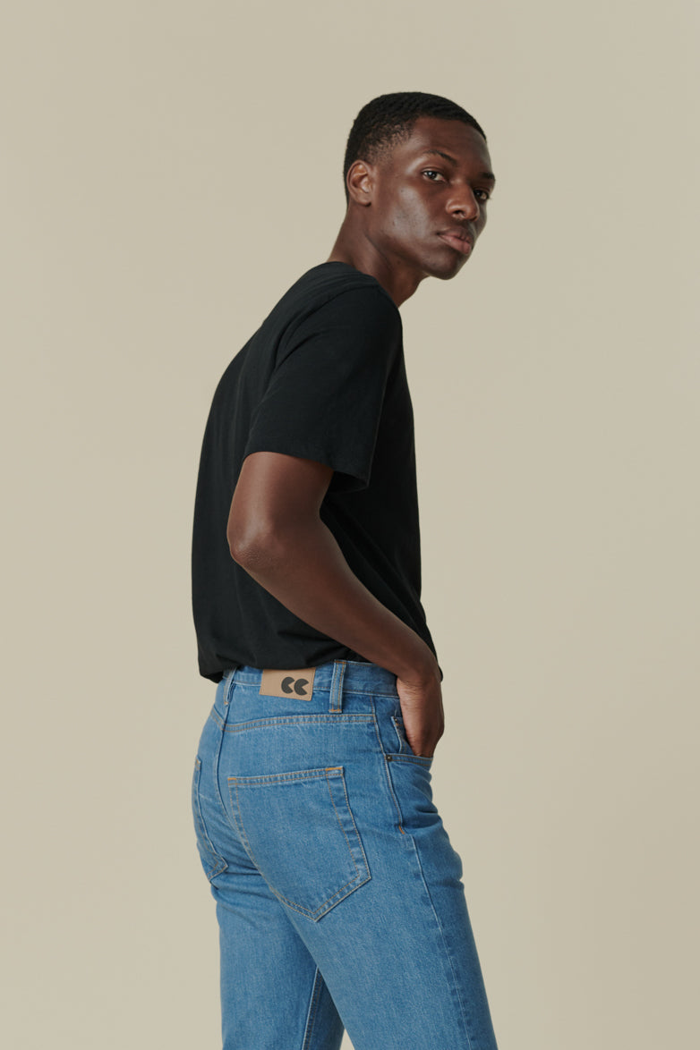 Thigh up image of male from the side looking back towards the camera, wearing short sleeve t shirt plastic free in black with straight cut selvedge jeans in fade