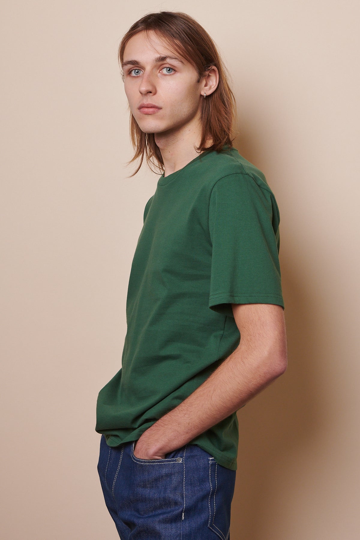 Side of male wearing short sleeve t shirt in bottle green with hands in front pockets of blue jeans. 