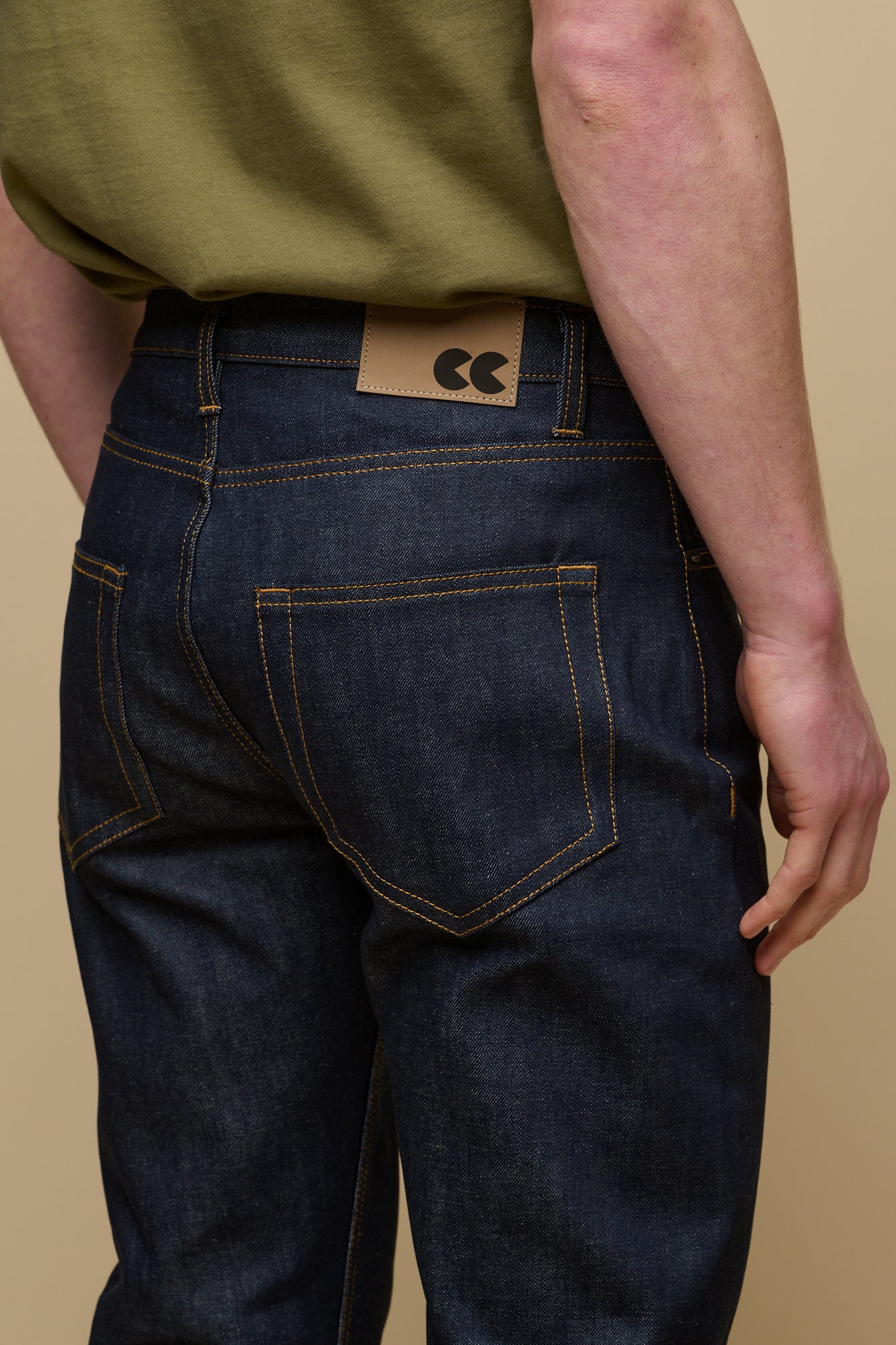 Close up detail shot of the back of men's straight cut selvedge jeans in indigo raw denim with tobacco contrast stitching.  Belt loops and CC logo jeans patch on the back of waistband with two back pockets.