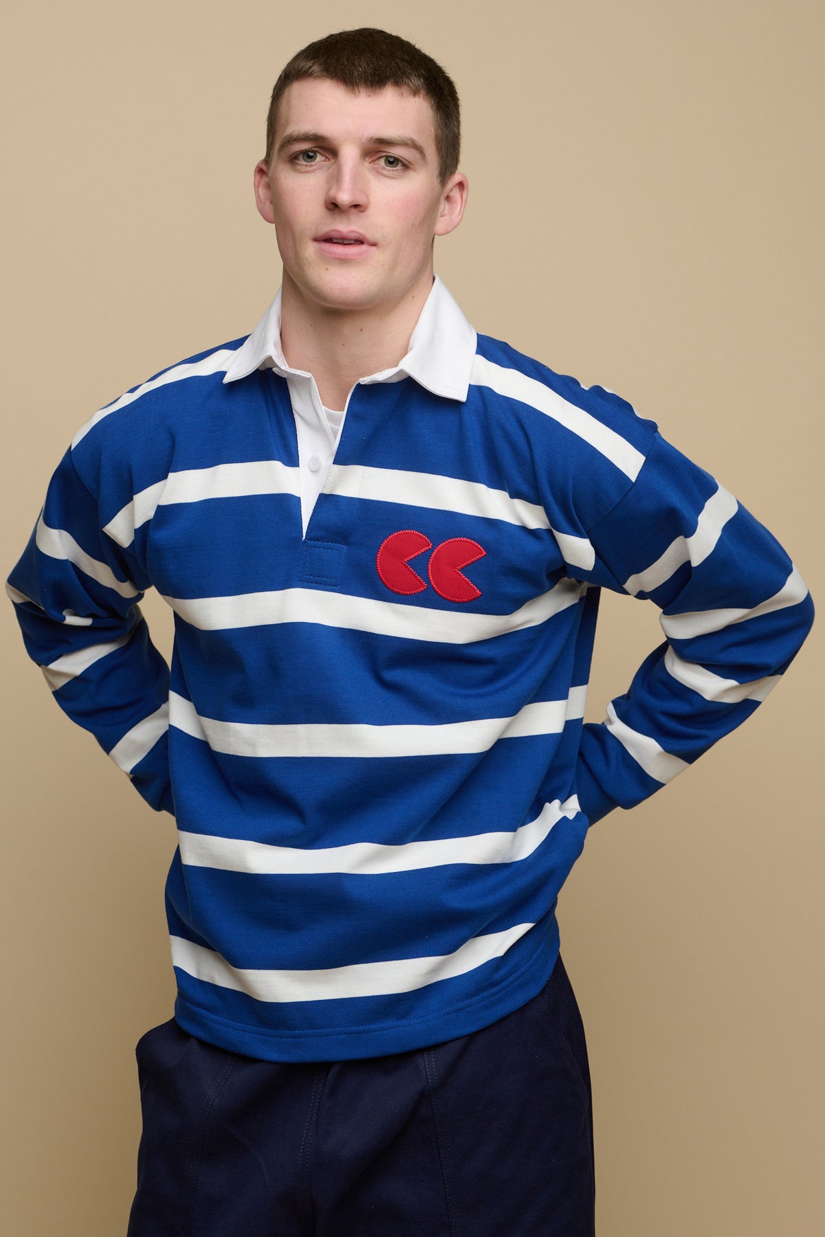 Image of brunet male from the thigh up with his hands on the back of his hips wearing blue and white stripe logo rugby shirt with red CC logo patch on chest, worn over crew neck white t shirt