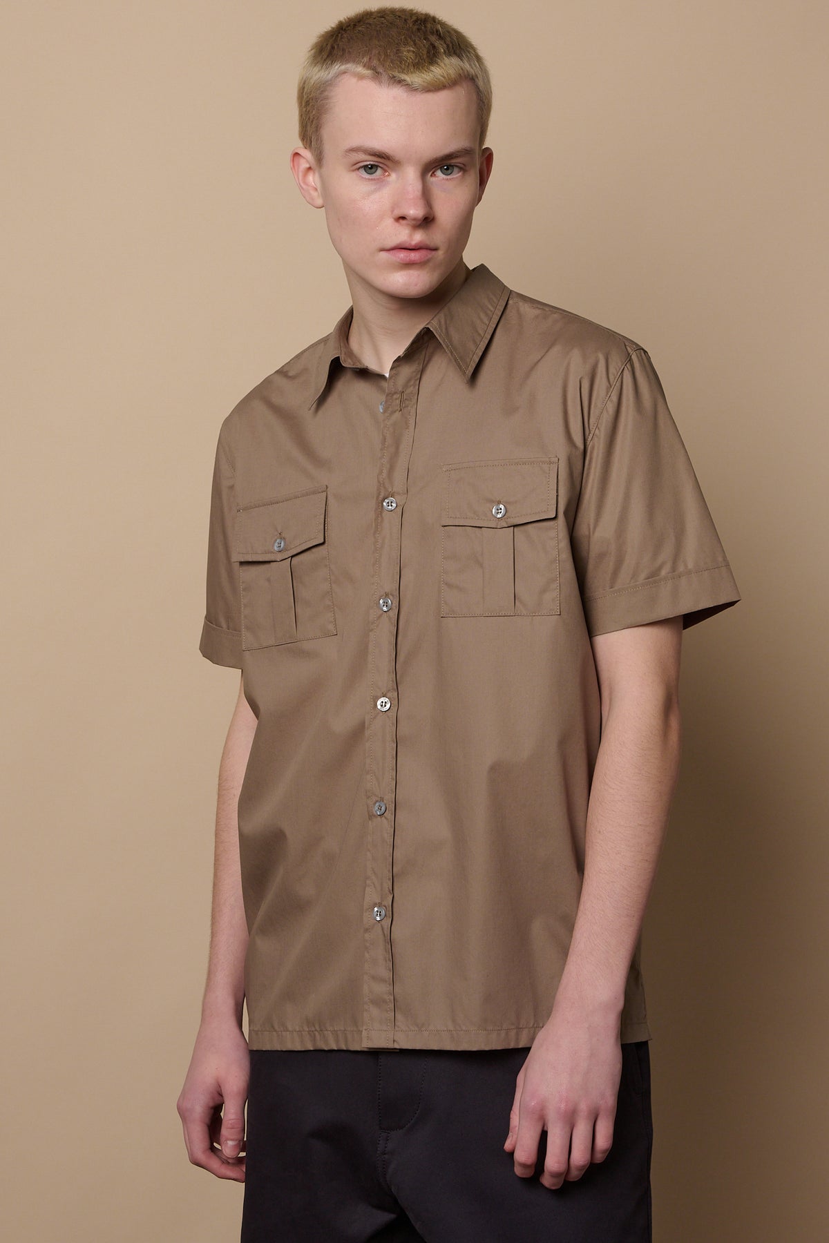 Thigh up image of white male wearing buttoned Tom short sleeve military shirt in khaki paired with chinos in navy 