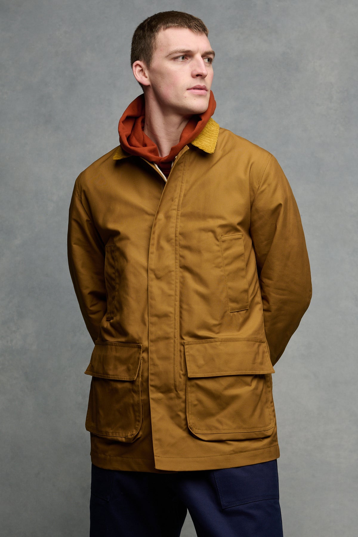 Thigh up image of brunet male wearing barn jacket in dark tan zipped up over hooded sweatshirt in cinnamon, paired with cameraman pants in navy