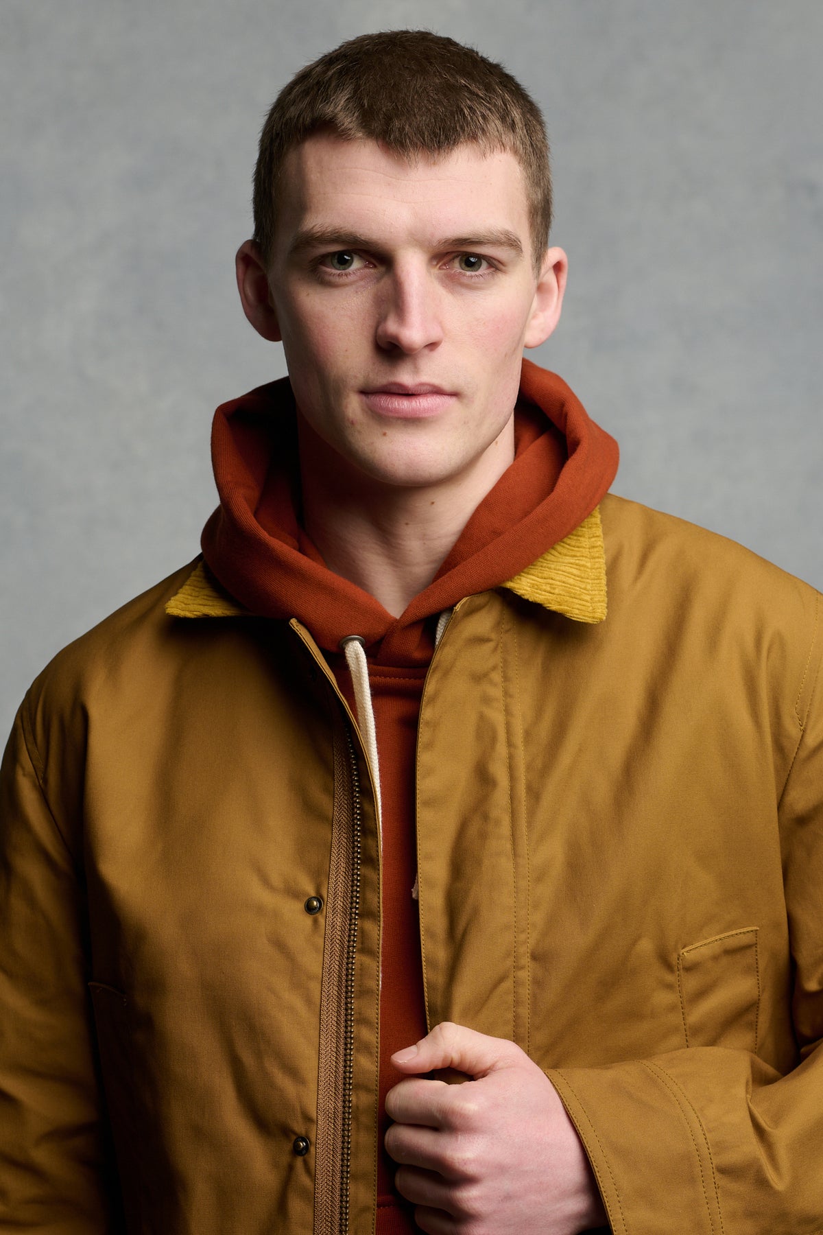 Portrait of male looking into the camera wearing men's barn jacket in dark tan with cord collar unzipped over cinnamon hooded sweatshirt.