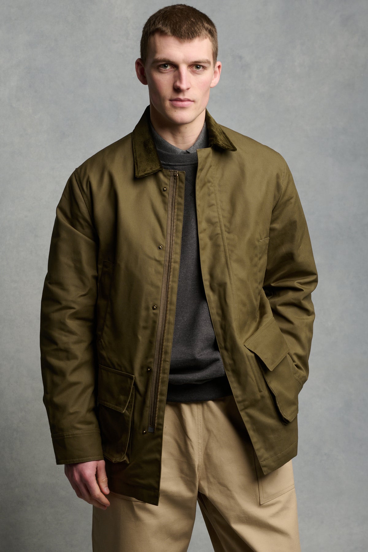Thigh up image of brunet male wearing unzipped barn jacket in olive worn over sweatshirt in charcoal paired with cameraman pant in putty