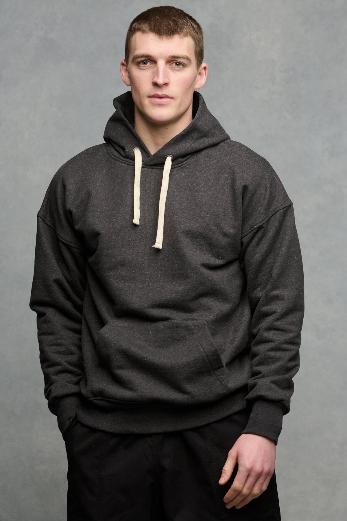 Thigh up front of brunet male wearing heritage hooded sweatshirt in charcoal paired with black cameraman pants