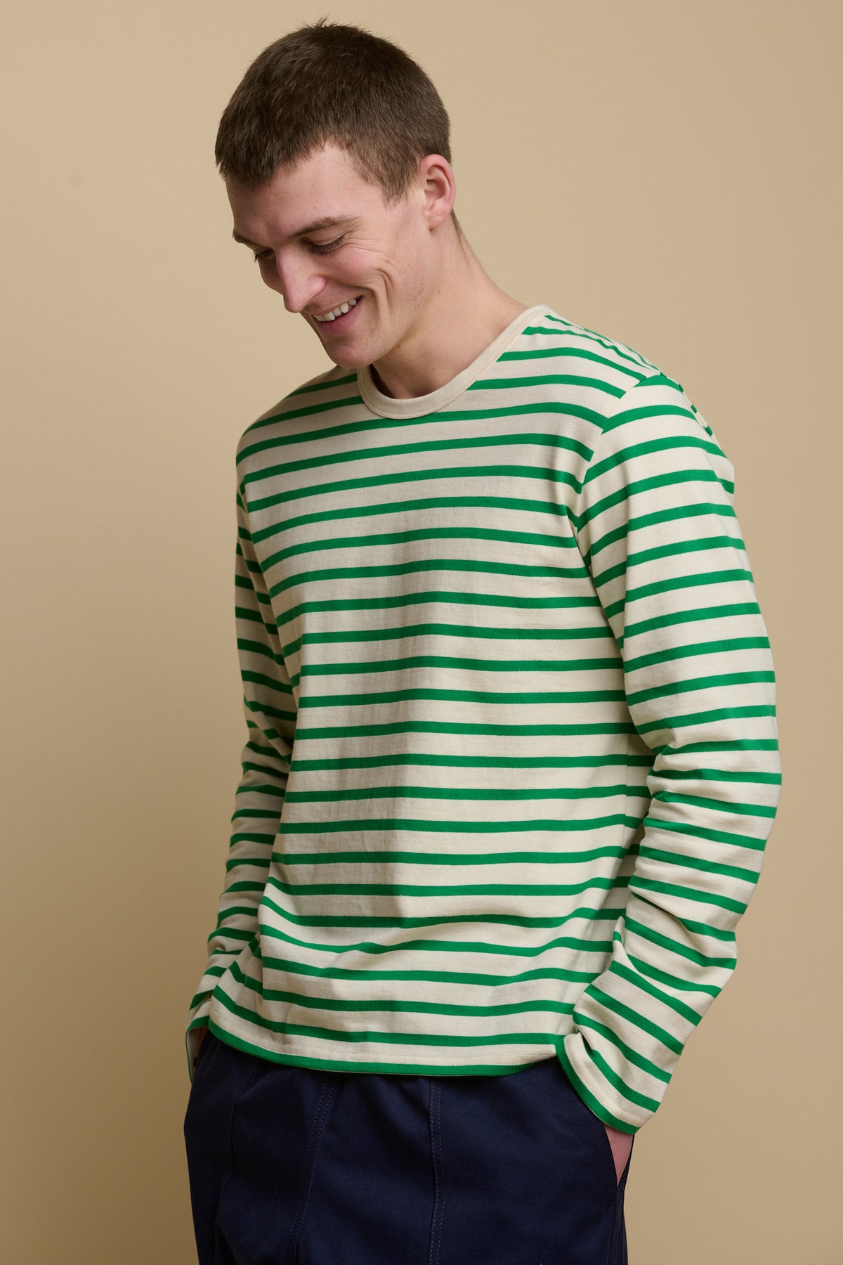 Thigh up image of smiley male looking towards the ground wearing Breton in ecru green with hands in the pockets of cameraman in navy