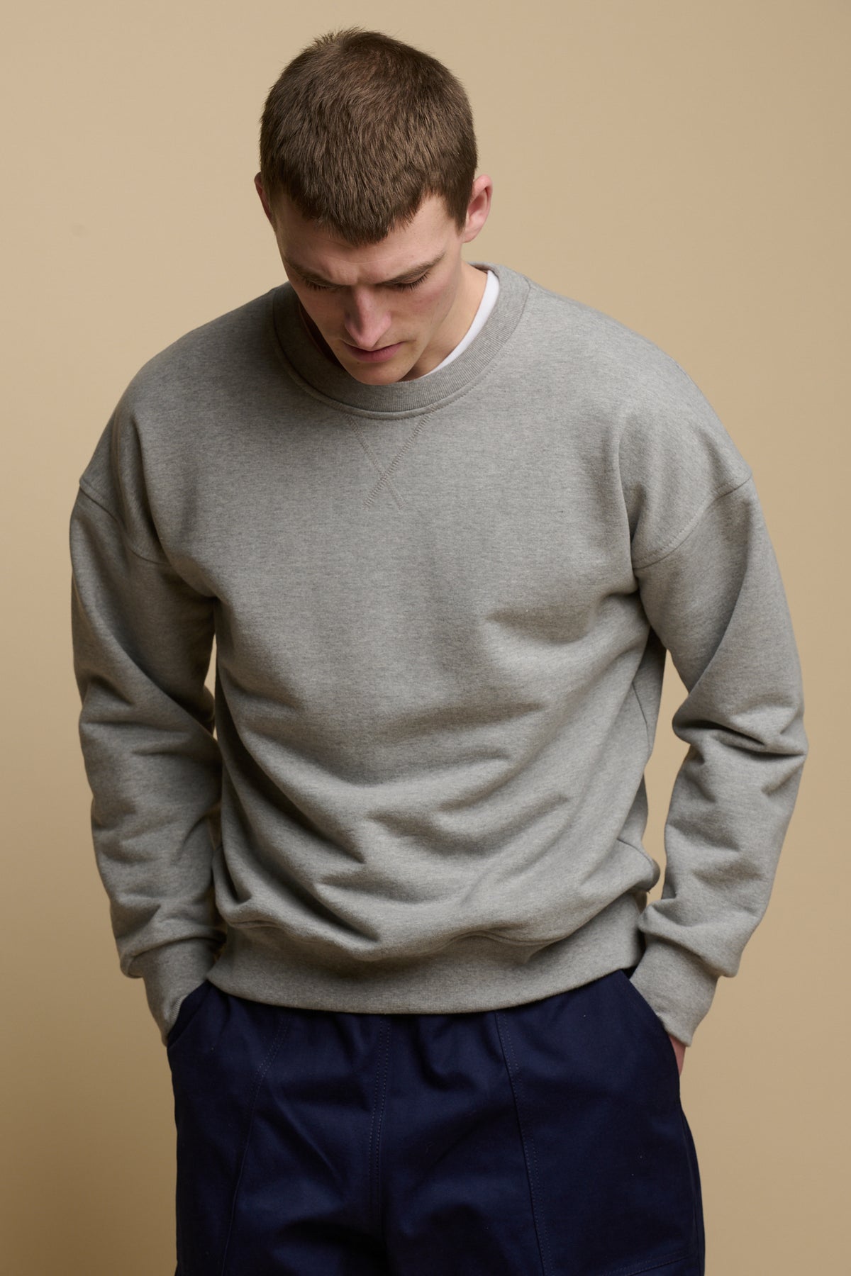 Thigh up front of brunet male looking down to the ground, wearing men's heritage drop shoulder sweatshirt in grey, worn over crew neck white t shirt paired with cameraman pants in navy