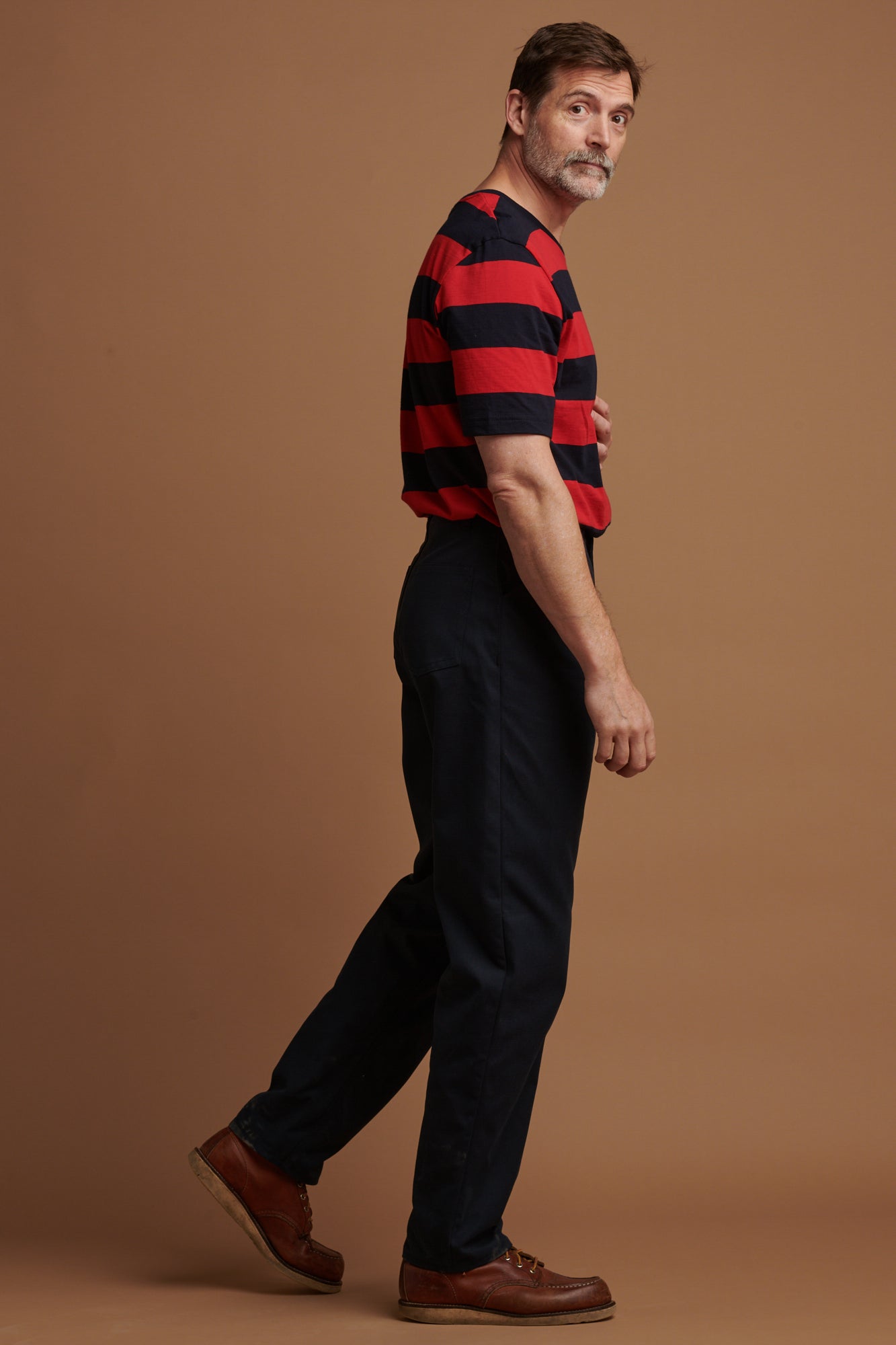 The right side of Patrick Grant looking back towards the camera wearing canvas chore trousers in navy paired with wide stripe t shirt in red/navy tucked in.