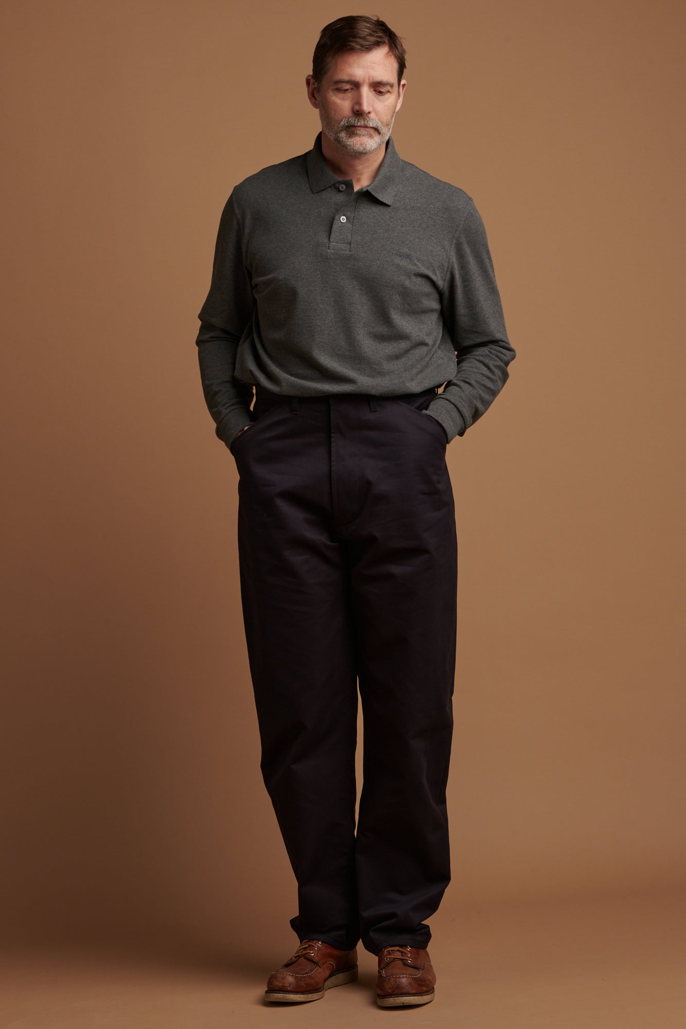 Patrick wearing cotton chore trousers with his hands in the front pockets paired with long sleeve polo shirt in grey