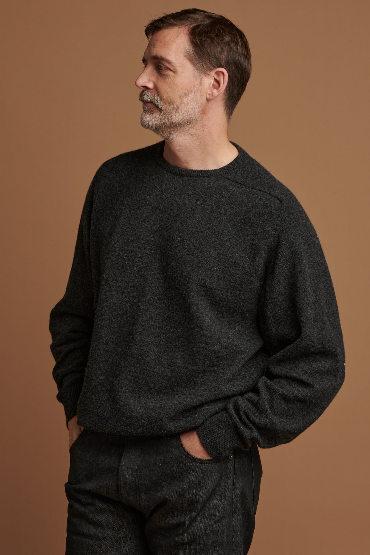 Patrick wearing lambswool saddle shoulder crew neck jumper in charcoal paired with loose fit button fly Arran jean in black