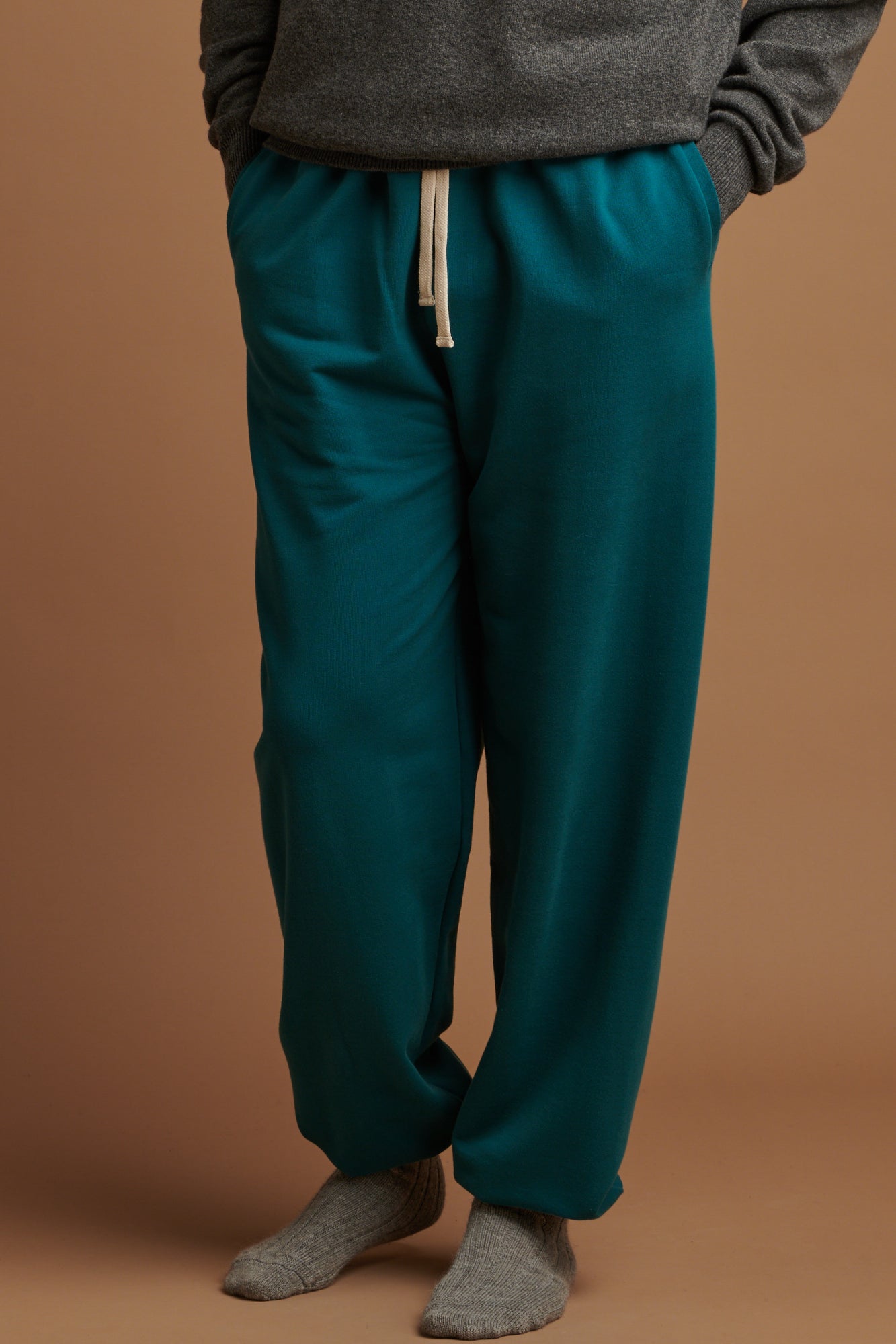 Waist down of the front of male wearing  men's sweatpants in teal, with hands in the front pockets and ecru drawcord on show, paired with alpaca bed socks