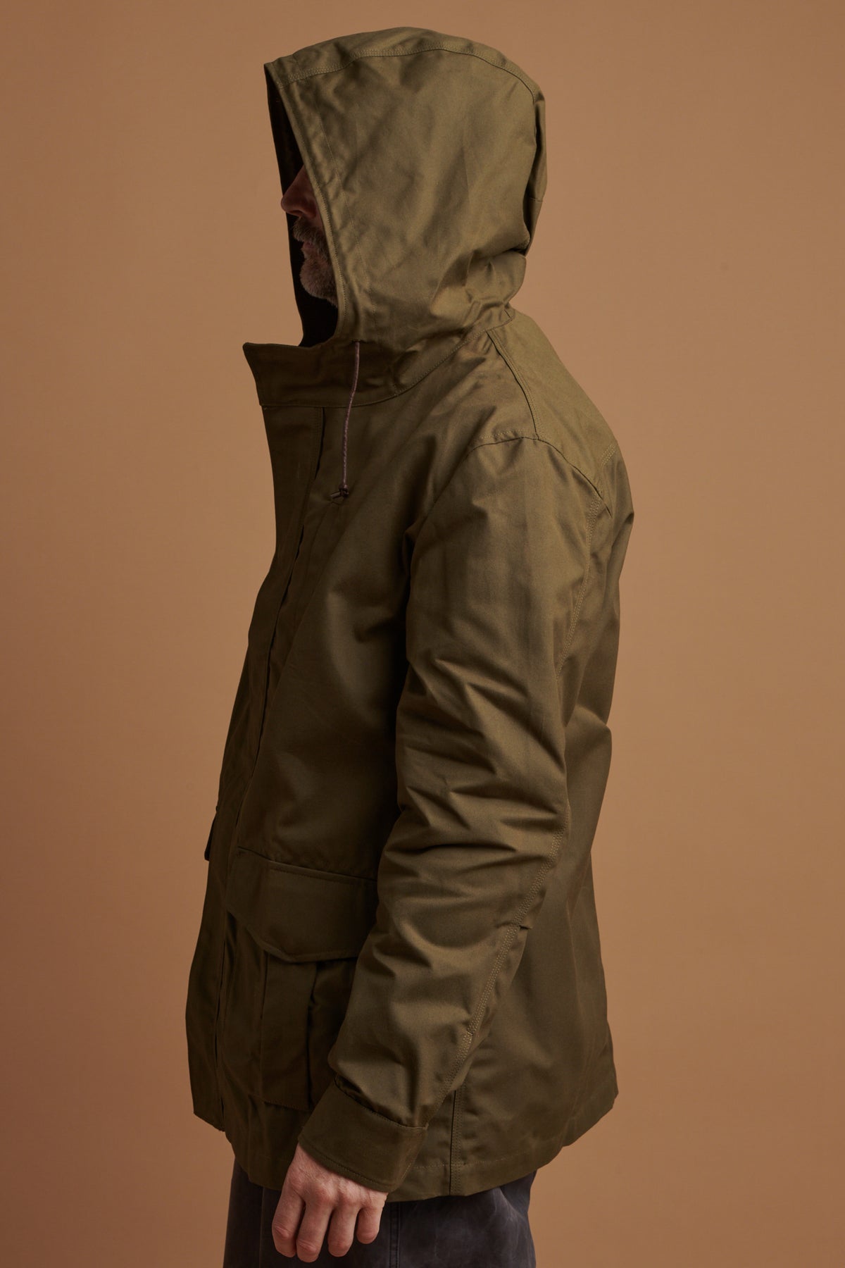 The side of Patrick wearing the waxed mountain parka in olive with the hood up 